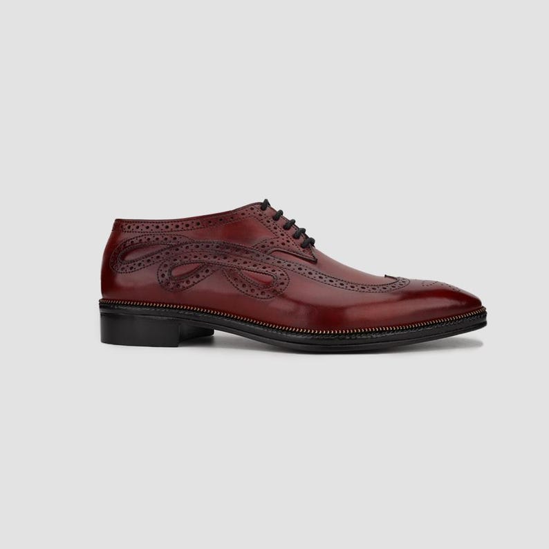 Puede incluir: Zapato de vestir de cuero burdeos con perforaciones decorativas y un dise&ntilde;o &uacute;nico en el lateral. El zapato tiene suela y cordones negros, con un tac&oacute;n bajo. Es un estilo cl&aacute;sico con un toque moderno.