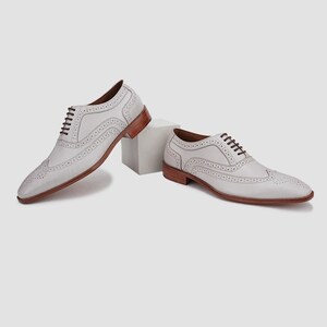 May include: Two white leather dress shoes with brown soles and heels. The shoes have decorative perforations and brown laces. One shoe is slightly elevated on a white cube.