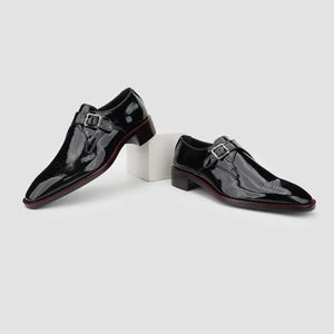 May include: A pair of black patent leather dress shoes with a single buckle closure. The shoes have a square toe and a low heel with a red accent along the sole. The shoes are displayed on a white surface.
