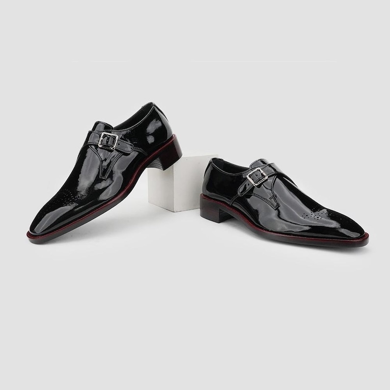 May include: A pair of black patent leather dress shoes with a single buckle closure. The shoes have a square toe and a low heel with a red accent along the sole. The shoes are displayed on a white surface.
