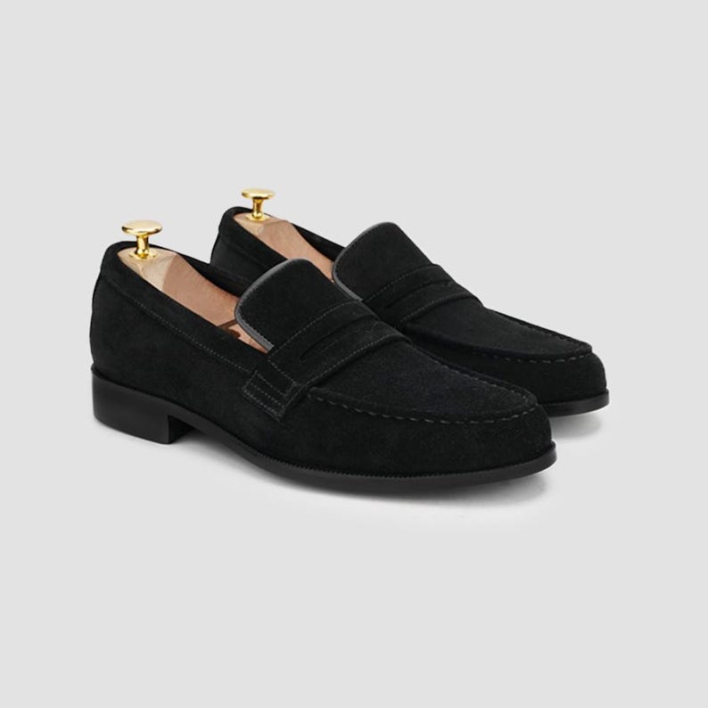 May include: A pair of black suede penny loafers with a classic design. The shoes feature a rounded toe, a strap across the vamp, and a low heel. Shoe trees are inserted inside the shoes to maintain their shape.