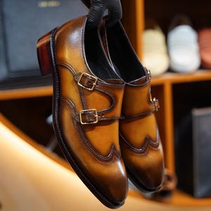 May include: A pair of brown leather dress shoes with a double monk strap design. The shoes have gold-coloured buckles and decorative stitching. The leather has a polished finish with a gradient of light to dark brown tones.