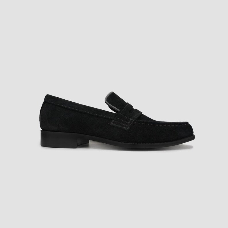 May include: Black suede penny loafers with a low heel. The shoes have a classic design with a strap across the vamp and a rounded toe. The loafers are a versatile footwear option.