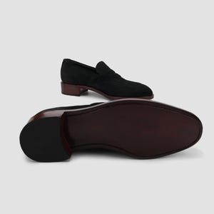 Puede incluir: Mocas&iacute;n de ante negro con suela y tac&oacute;n marr&oacute;n oscuro. El zapato presenta un dise&ntilde;o cl&aacute;sico sin cordones con punta redondeada. El tac&oacute;n mide aproximadamente 2,5 cm.
