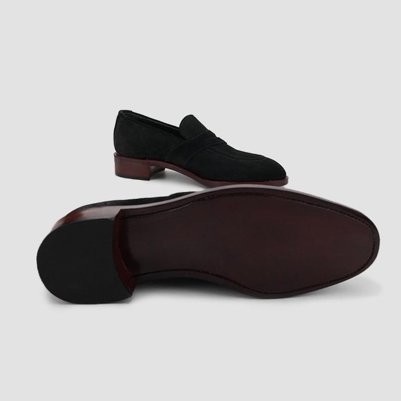 Puede incluir: Mocas&iacute;n de ante negro con suela y tac&oacute;n marr&oacute;n oscuro. El zapato presenta un dise&ntilde;o cl&aacute;sico sin cordones con punta redondeada. El tac&oacute;n mide aproximadamente 2,5 cm.