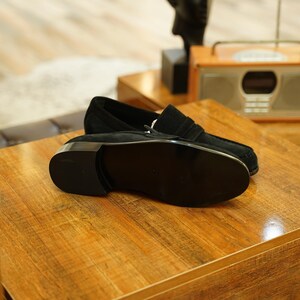 May include: A pair of black suede loafers with a silver-toned buckle, resting on a wooden surface. The shoes have a classic design with a rounded toe and a low heel. The background includes a vintage-style radio.