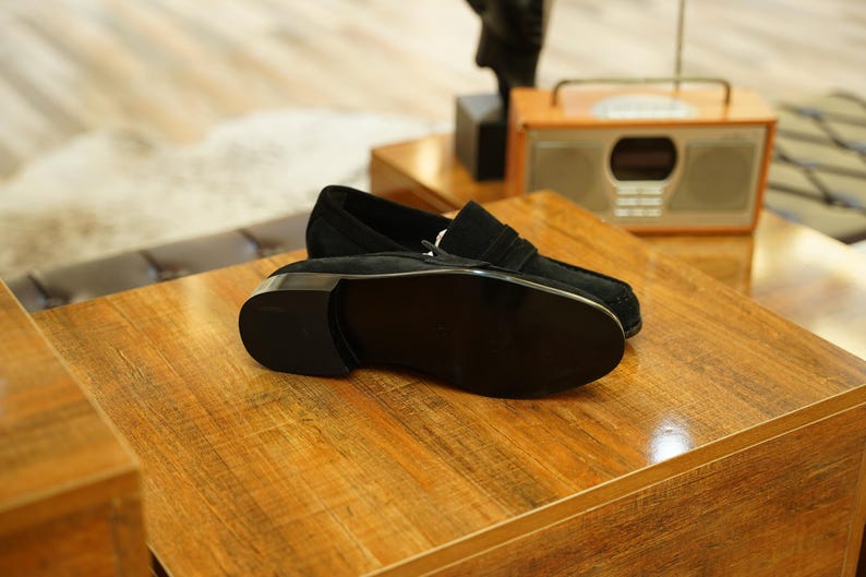 May include: A pair of black suede loafers with a silver-toned buckle, resting on a wooden surface. The shoes have a classic design with a rounded toe and a low heel. The background includes a vintage-style radio.