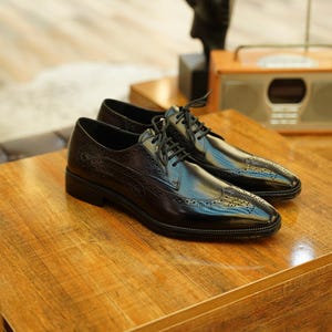 Puede incluir: Un par de zapatos de vestir negros de cuero con detalles de ala y cierre de cordones. Los zapatos se exhiben sobre una superficie de madera, mostrando su acabado brillante y su intrincado dise&ntilde;o. Parte de una colecci&oacute;n de calzado para hombre.