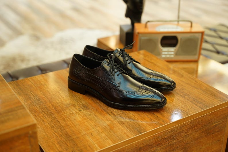 Puede incluir: Un par de zapatos de vestir negros de cuero con detalles de ala y cierre de cordones. Los zapatos se exhiben sobre una superficie de madera, mostrando su acabado brillante y su intrincado dise&ntilde;o. Parte de una colecci&oacute;n de calzado para hombre.