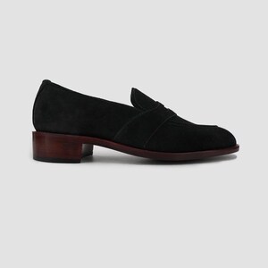 Puede incluir: Mocas&iacute;n de ante negro con tac&oacute;n bajo marr&oacute;n oscuro. El zapato presenta un dise&ntilde;o cl&aacute;sico con puntera redondeada y una correa decorativa en el empeine. La suela es de color marr&oacute;n oscuro a juego.