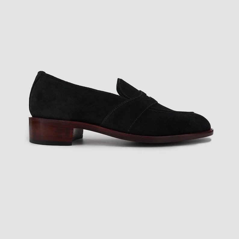 Puede incluir: Mocas&iacute;n de ante negro con tac&oacute;n bajo marr&oacute;n oscuro. El zapato presenta un dise&ntilde;o cl&aacute;sico con puntera redondeada y una correa decorativa en el empeine. La suela es de color marr&oacute;n oscuro a juego.
