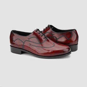 May include: A pair of burgundy leather dress shoes with black laces. The shoes have a glossy finish and black stitching details. The soles and heels are black. The shoes are displayed on a light gray background.