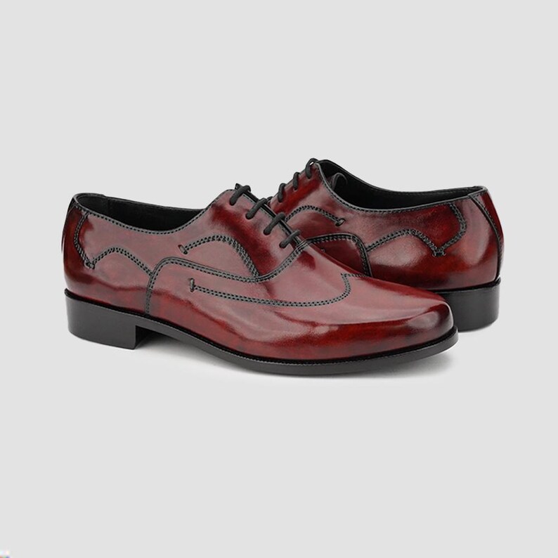 May include: A pair of burgundy leather dress shoes with black laces. The shoes have a glossy finish and black stitching details. The soles and heels are black. The shoes are displayed on a light gray background.