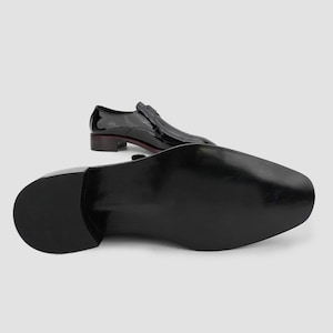 May include: Black patent leather dress shoes with a low heel. One shoe is positioned with the sole facing the viewer, while the other is in the background, showing the top of the shoe. The shoes have a sleek, polished appearance.