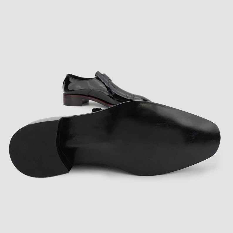 May include: Black patent leather dress shoes with a low heel. One shoe is positioned with the sole facing the viewer, while the other is in the background, showing the top of the shoe. The shoes have a sleek, polished appearance.