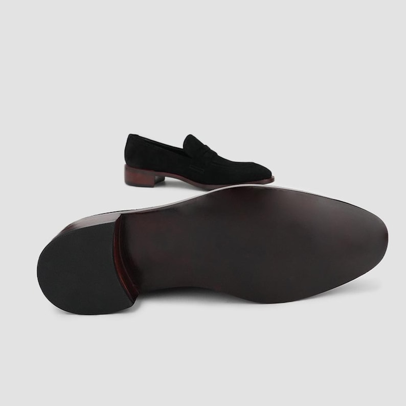 May include: Black suede loafer with a low heel and a dark brown sole. The shoe is displayed with the sole facing up, showcasing the craftsmanship and design of the footwear. A second shoe is positioned above, offering a view of the upper.
