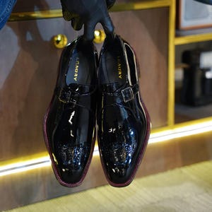 May include: A pair of glossy black leather dress shoes with a buckle closure. The shoes have a textured toe cap and a red trim along the sole. The word "LEAGURY" is printed on the insole.