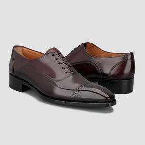 May include: A pair of dark burgundy leather oxford shoes featuring a polished finish, a cap toe with subtle decorative perforations, a low stacked heel, and matching dark laces. The shoes have a sleek, structured silhouette with clean lines.