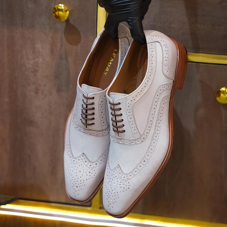 May include: A pair of white leather dress shoes with brown laces and soles. The shoes have a wingtip design with decorative perforations and are held up by a gloved hand. The brand name "LEMORY" is visible inside the shoe.