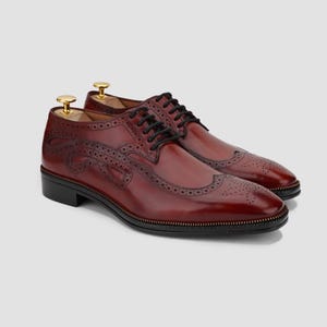Puede incluir: Zapatos de vestir de cuero burdeos con cordones negros y dise&ntilde;o decorativo lateral. Los zapatos presentan un dise&ntilde;o brogue cl&aacute;sico con perforaciones y suela negra. Se insertan hormas. Los zapatos miden aproximadamente 30 cm de largo.