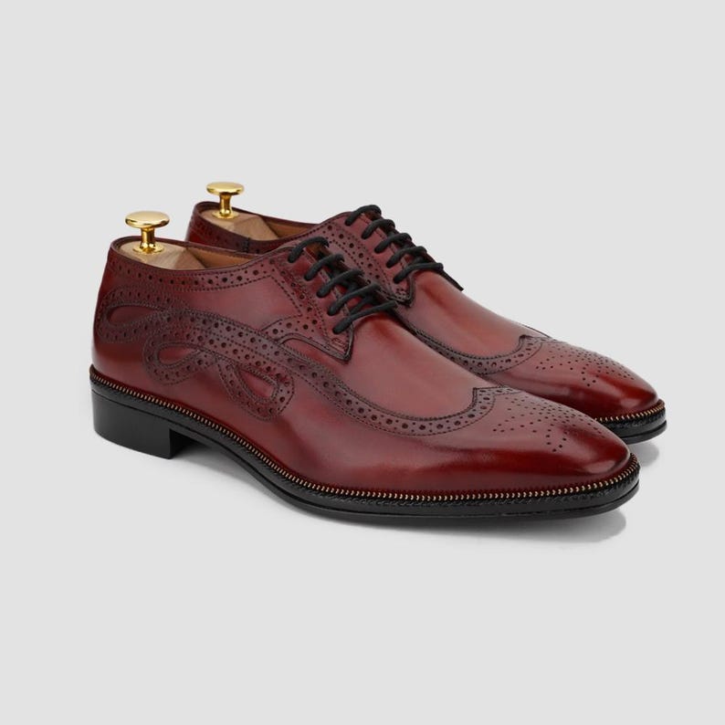 Puede incluir: Zapatos de vestir de cuero burdeos con cordones negros y dise&ntilde;o decorativo lateral. Los zapatos presentan un dise&ntilde;o brogue cl&aacute;sico con perforaciones y suela negra. Se insertan hormas. Los zapatos miden aproximadamente 30 cm de largo.