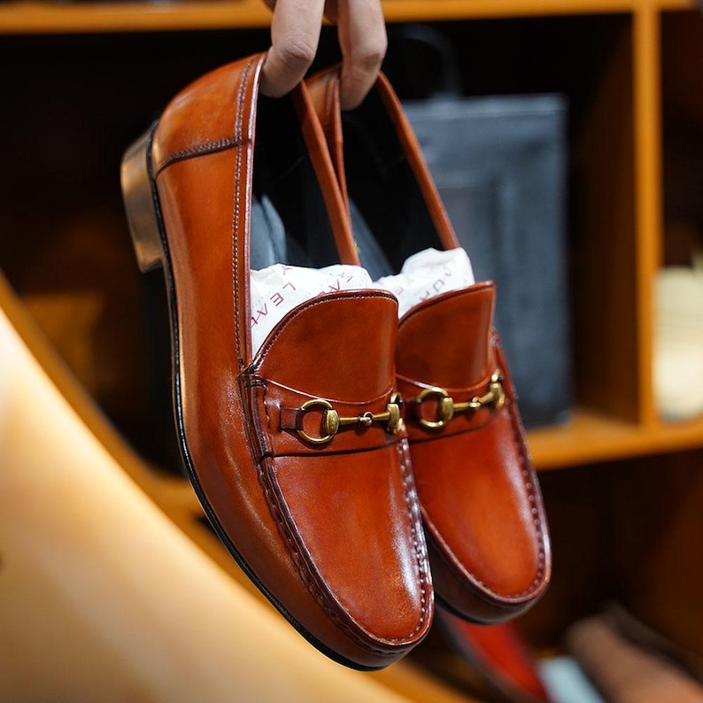 May include: A pair of brown leather loafers with gold-colored metal accents. The shoes are held up, showcasing their polished finish and classic design. The loafers have a slip-on style with a decorative buckle.