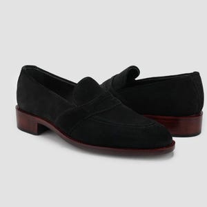 Puede incluir: Un par de mocasines negros de ante con tac&oacute;n bajo y suela marr&oacute;n oscuro. Los zapatos tienen un dise&ntilde;o cl&aacute;sico con una pala cosida y una correa en la parte superior. Los tacones miden aproximadamente 2,5 cm de alto.