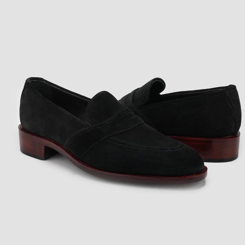 Puede incluir: Un par de mocasines negros de ante con tac&oacute;n bajo y suela marr&oacute;n oscuro. Los zapatos tienen un dise&ntilde;o cl&aacute;sico con una pala cosida y una correa en la parte superior. Los tacones miden aproximadamente 2,5 cm de alto.