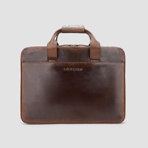 May include: A brown leather briefcase with a top handle and a shoulder strap. The briefcase has a rectangular shape with a zipper closure and the brand name "LEAGUV" embossed on the front. The leather has a polished finish.