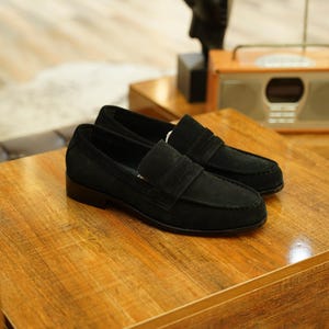 May include: A pair of black suede loafers with a classic design, featuring a strap across the vamp. The shoes are displayed on a wooden surface, showcasing their sleek and sophisticated style. These loafers are a timeless footwear choice.