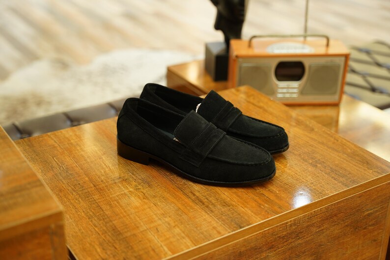 May include: A pair of black suede loafers with a classic design, featuring a strap across the vamp. The shoes are displayed on a wooden surface, showcasing their sleek and sophisticated style. These loafers are a timeless footwear choice.