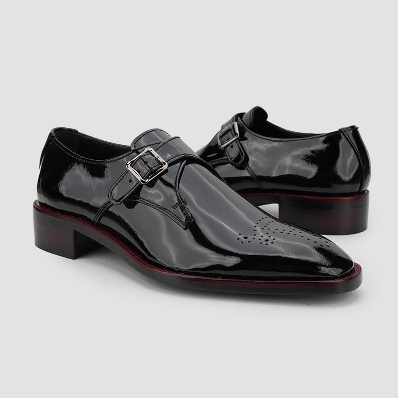 May include: Pair of black patent leather dress shoes with a buckle closure. The shoes have a low heel and a decorative perforated design on the toe. The soles have a red trim.