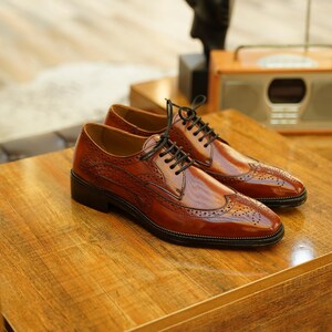 Puede incluir: Un par de zapatos de vestir de cuero marr&oacute;n con cordones negros, exhibidos sobre una superficie de madera. Los zapatos tienen un dise&ntilde;o de ala con perforaciones decorativas. El fondo incluye una radio vintage y otra decoraci&oacute;n.