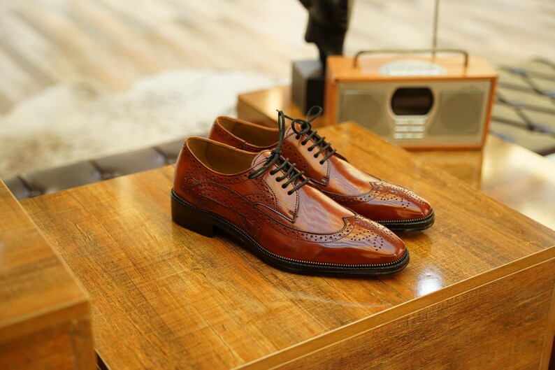 Puede incluir: Un par de zapatos de vestir de cuero marr&oacute;n con cordones negros, exhibidos sobre una superficie de madera. Los zapatos tienen un dise&ntilde;o de ala con perforaciones decorativas. El fondo incluye una radio vintage y otra decoraci&oacute;n.