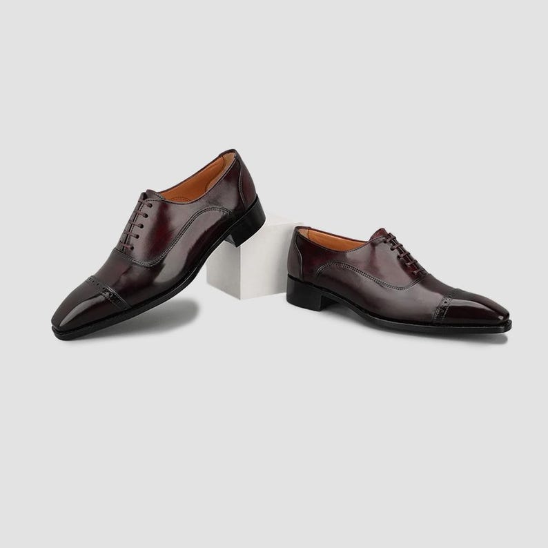 May include: A pair of dark burgundy leather oxford shoes with a cap toe design, thin matching laces, and a low stacked heel. The shoes feature a polished finish, a rounded toe box, and a structured silhouette with visible stitching along the seams.