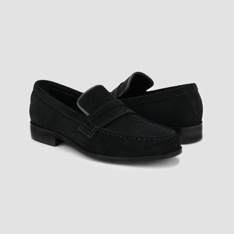 May include: A pair of black suede penny loafers. The shoes have a classic design with a rounded toe, a strap across the vamp, and a low heel. The stitching is visible, and the shoes are set against a neutral background.