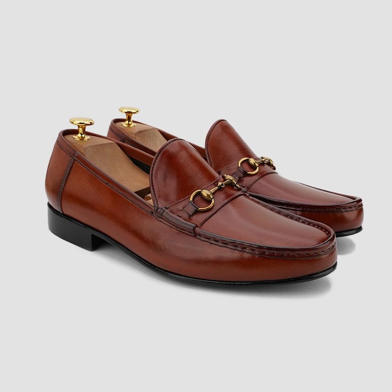 May include: A pair of brown leather loafers with gold-colored metal accents. The shoes have a classic design with a rounded toe and a slip-on style. The loafers are displayed with wooden shoe trees inside.