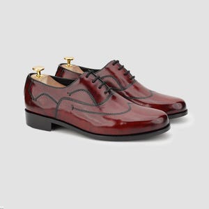May include: A pair of burgundy leather Oxford shoes with black stitching and laces. The shoes have a glossy finish and are displayed with wooden shoe trees. The classic design features a wingtip style and a low heel.