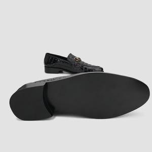 May include: Black leather loafers with a crocodile texture. The shoes have a gold-colored metal buckle detail. One shoe is positioned with the sole facing the viewer, and the other is in the background.