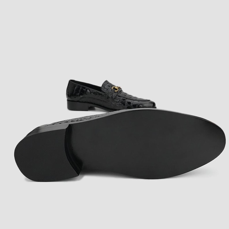 May include: Black leather loafers with a crocodile texture. The shoes have a gold-colored metal buckle detail. One shoe is positioned with the sole facing the viewer, and the other is in the background.