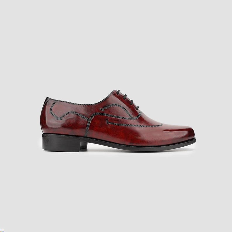 May include: A single, polished burgundy leather dress shoe with black stitching and laces. The shoe features a classic Oxford design with a low heel and a sleek, pointed toe. The shoe is designed for formal occasions.