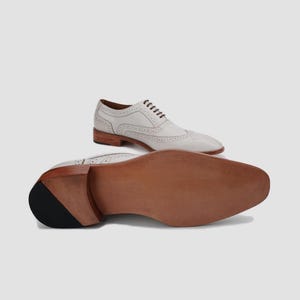 May include: A pair of white leather wingtip dress shoes with brown soles and laces. The shoes have decorative perforations and a classic, formal design. The image shows the shoes from two different angles, one from the side and the other from the sole.