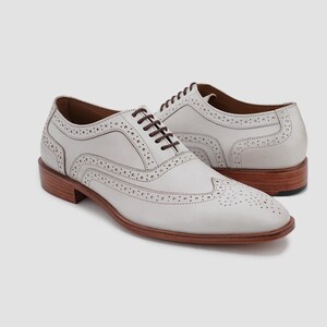 May include: A pair of white leather wingtip dress shoes with brown laces and a brown sole. The shoes have decorative perforations and are displayed on a neutral background.