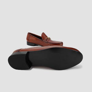 May include: Brown leather loafers with a gold-colored metal horsebit detail. The shoes have a classic design with a rounded toe and a low heel. The sole is black. One shoe is upright, and the other is lying on its side.