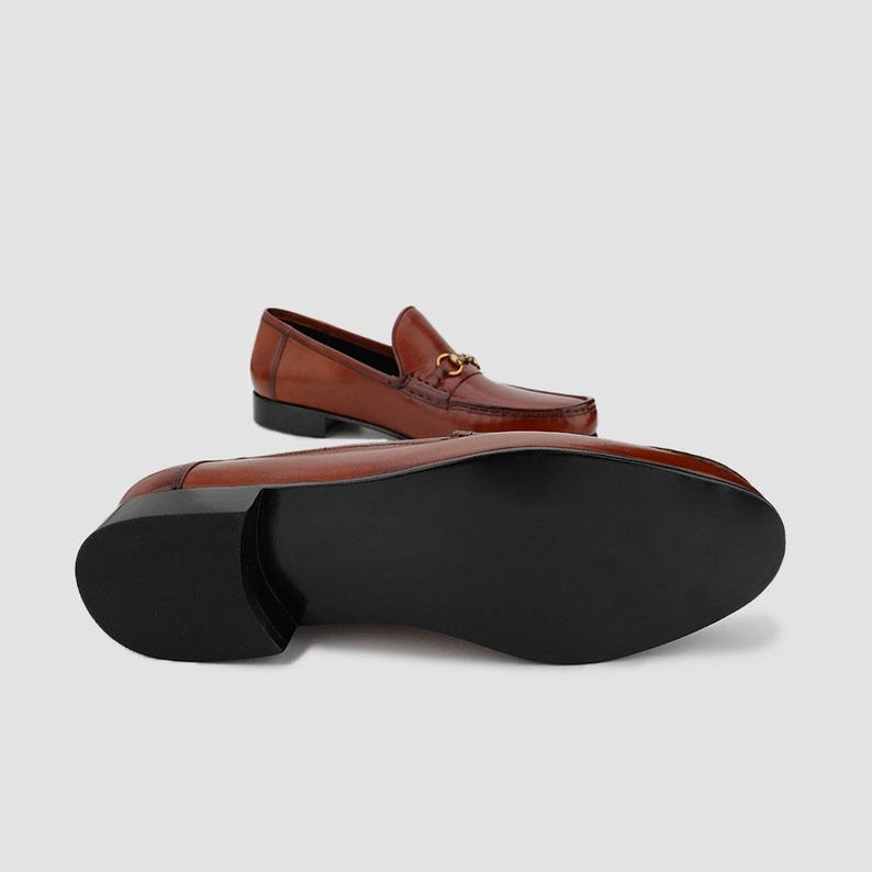 May include: Brown leather loafers with a gold-colored metal horsebit detail. The shoes have a classic design with a rounded toe and a low heel. The sole is black. One shoe is upright, and the other is lying on its side.