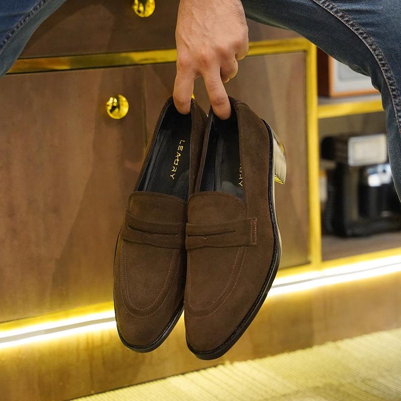 May include: A pair of brown suede loafers with a classic design. The shoes have a rounded toe, a strap across the vamp, and a stacked heel. The brand name "LE MDAY" is visible on the insole.
