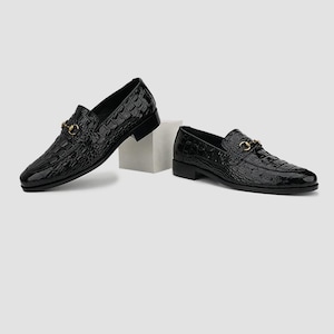 May include: A pair of black leather loafers with a crocodile texture. Each shoe features a gold-colored metal buckle detail. The shoes are displayed on a white background, showcasing their glossy finish and classic design.