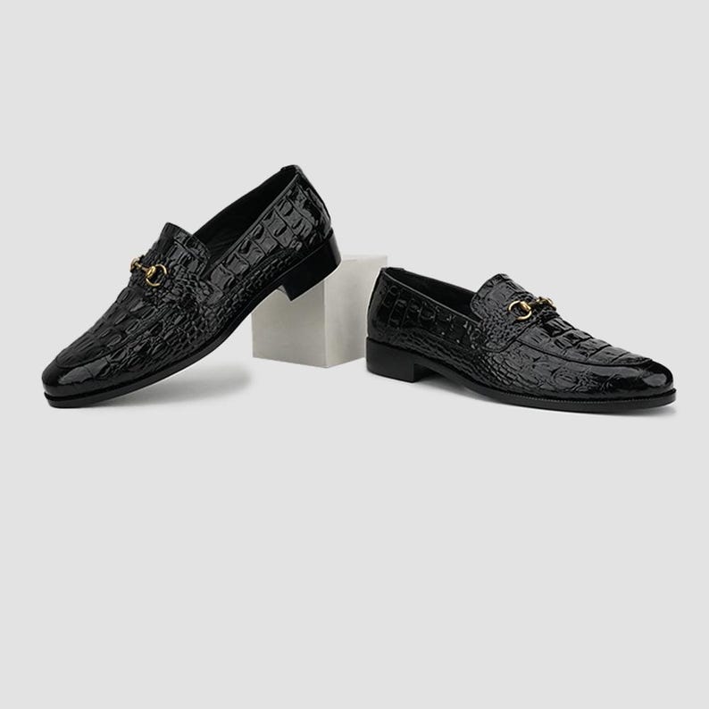 May include: A pair of black leather loafers with a crocodile texture. Each shoe features a gold-colored metal buckle detail. The shoes are displayed on a white background, showcasing their glossy finish and classic design.