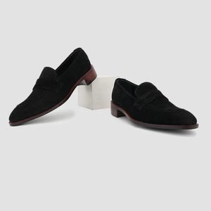 May include: A pair of black suede penny loafers with a dark brown heel and sole. The shoes are displayed on a white surface, showcasing their classic design and elegant silhouette. The loafers have a slip-on style.