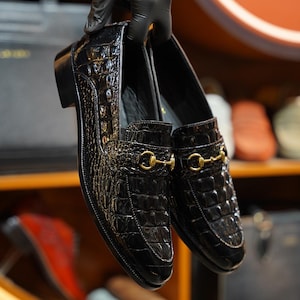 May include: A pair of black leather loafers with a crocodile skin texture. The shoes have a gold-colored metal bit detail on the vamp. The shoes are held up by a gloved hand.